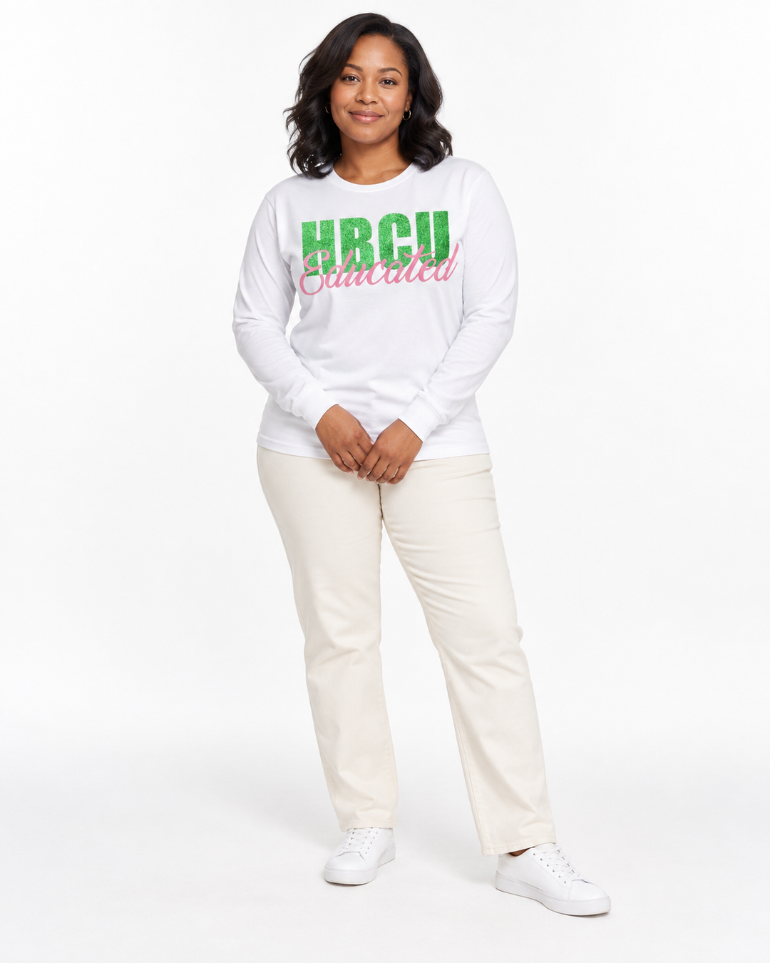 Model with hands loosely clasped wearing a white long-sleeve HBCU Educated Tee with green glitter lettering and matte pink script.