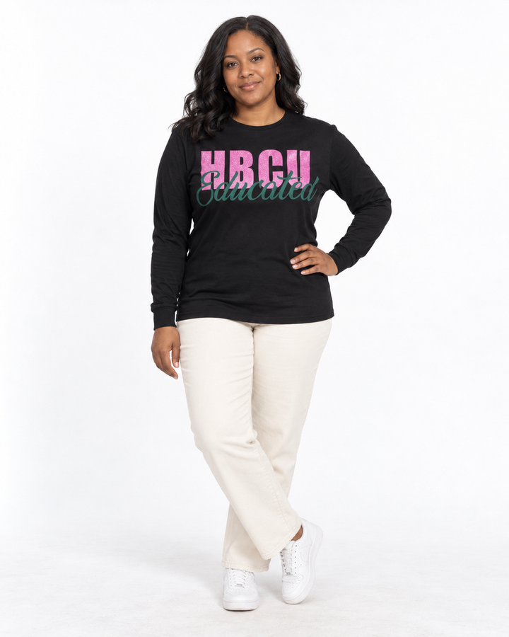 Model in a crossed-ankle hand-on-hip pose wearing a black long-sleeve HBCU Educated Tee with pink glitter lettering and matte green script.